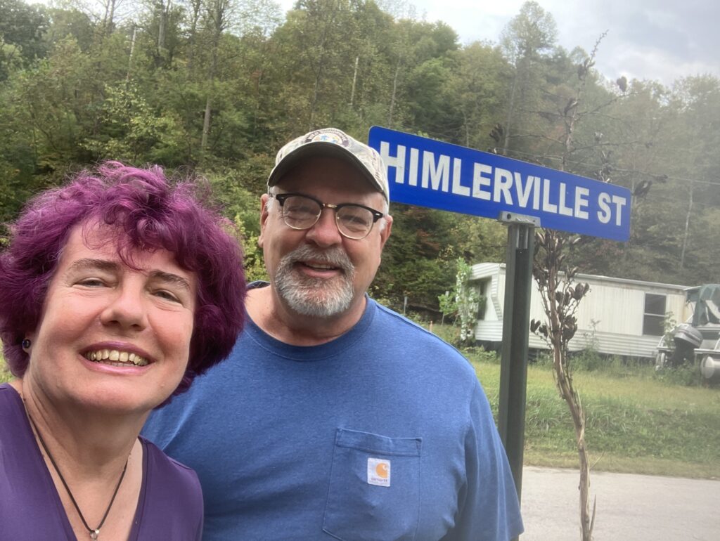 Two people smiling with a "HIMLERVILLE ST" street sign and wooded area in the background.
