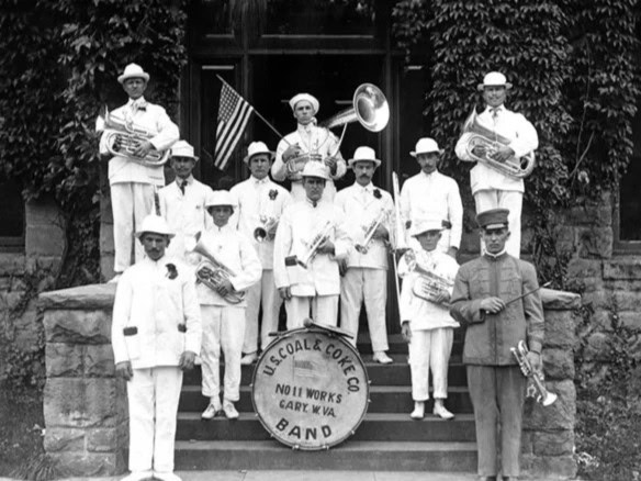 A historical marching band in white uniforms poses with instruments on stone steps.
