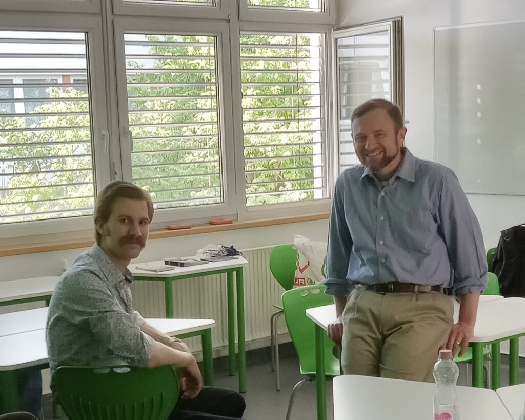People in a classroom with green chairs and white desks, near large windows with outside greenery.