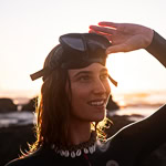 Person in a wetsuit with goggles on head, smiling and shielding eyes from sunlight.