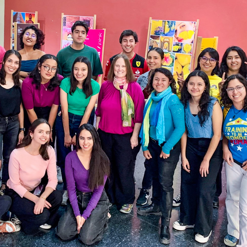 A group of fourteen people posing indoors against a red wall with colorful posters.