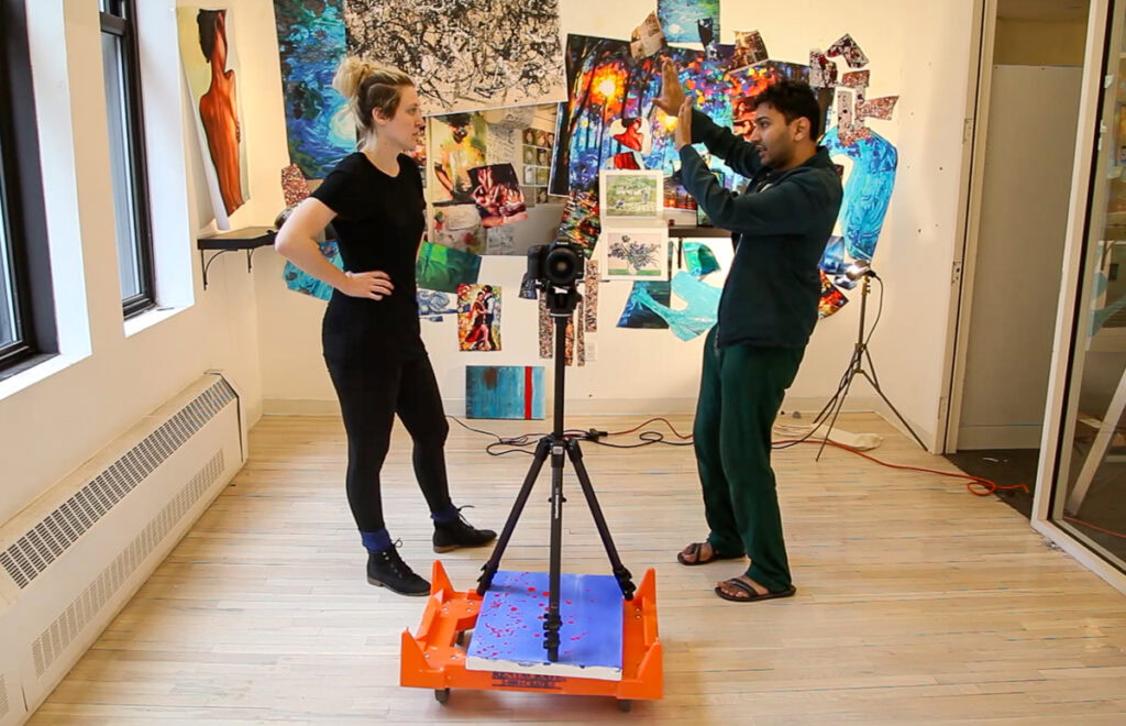 Two people in an art studio with a camera on a tripod surrounded by colorful artwork.