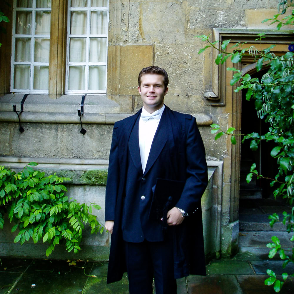 McLean in formal academic attire standing in front of an old stone building with windows and an arched doorway.