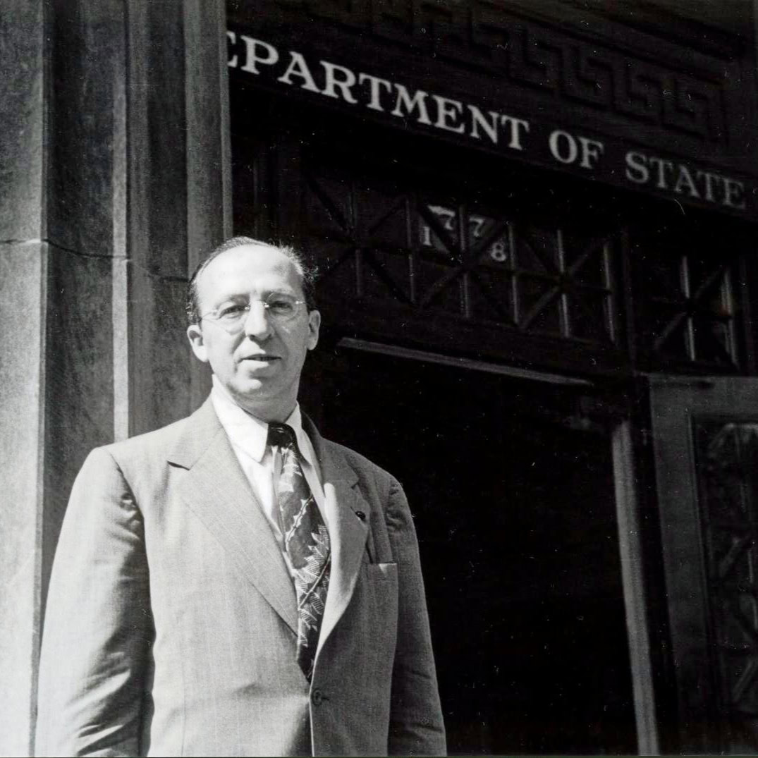 Copland wearing a suit and tie and  standing in front of doorway with sign above it reading U.S. Department of State, 