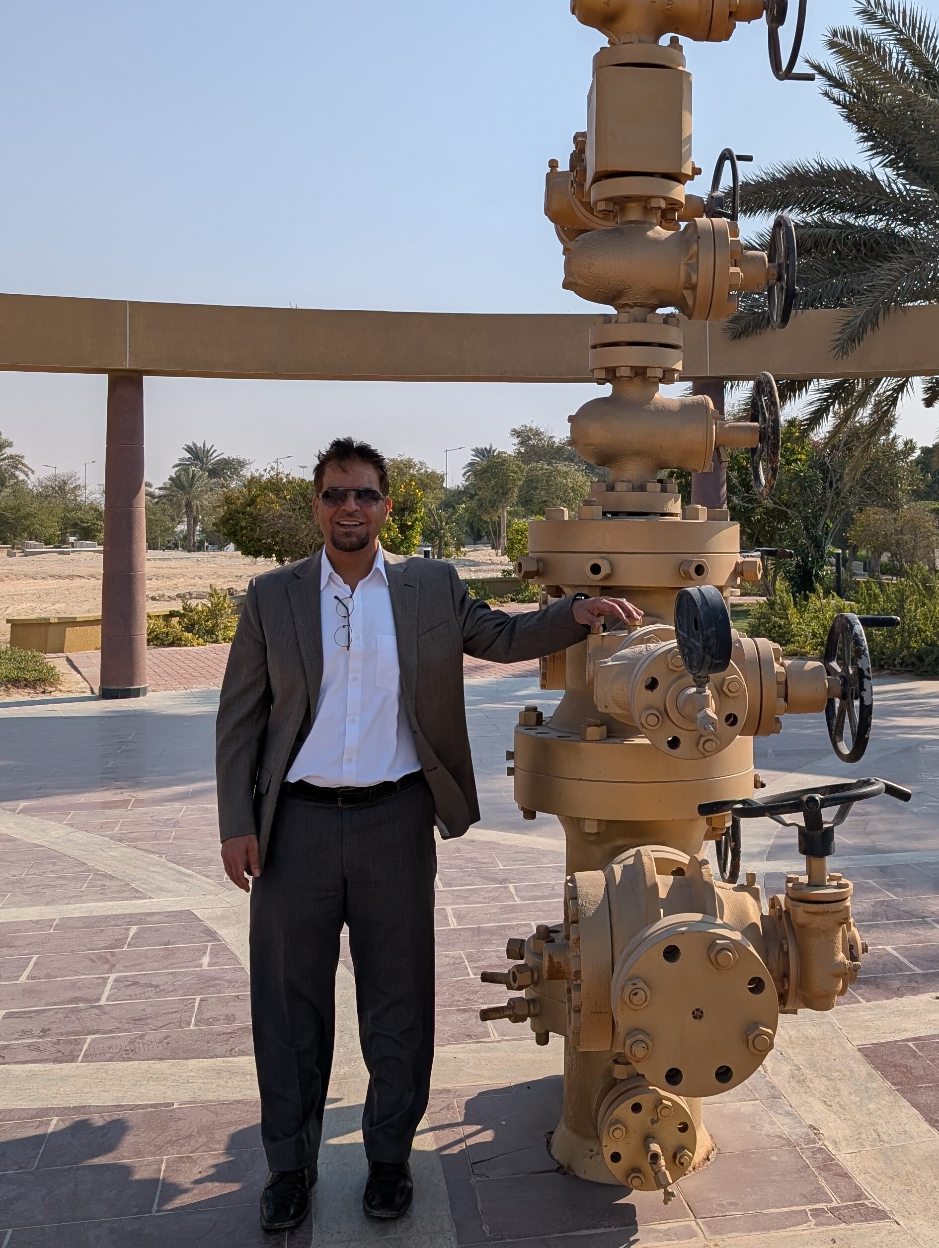 Alshami standing next to oil well equipment, outdoors with palm trees.

