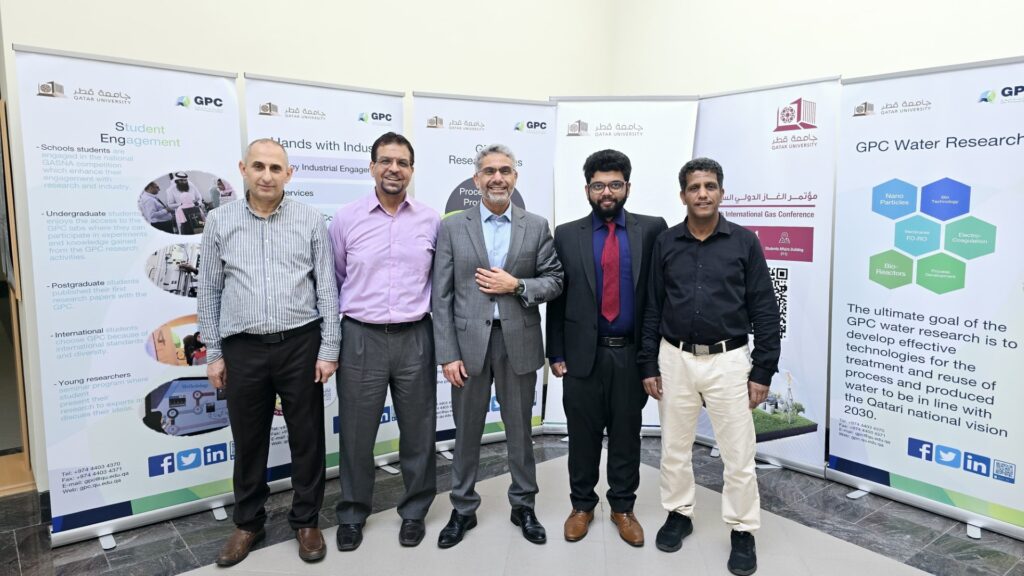 Alshami standing with four other men wearing business attire standing in front of several large conference banners describing Qatar University and water research