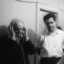 Copeland standing holding headphone speakers over his ears, listening to music with Amar Bose, music system inventor, looking on from his left.