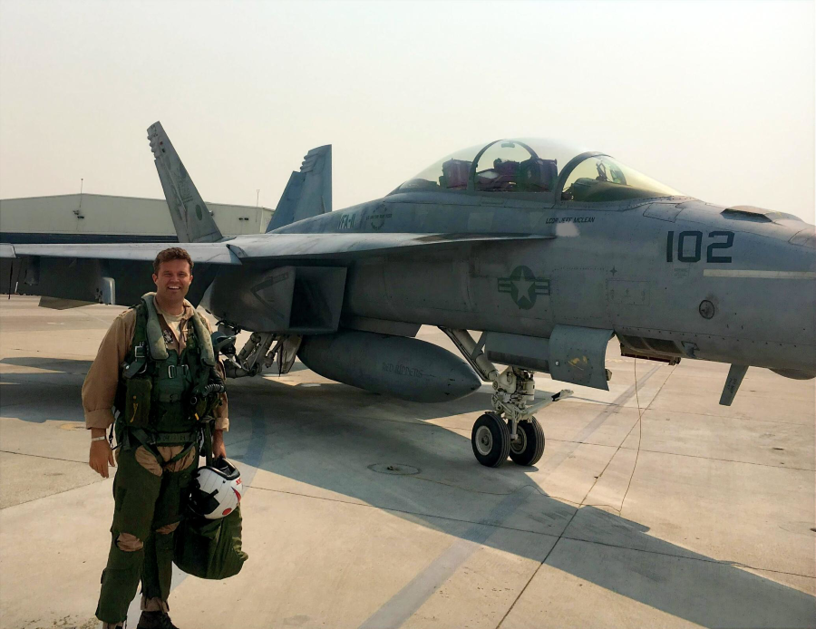McLean, a pilot stands in front of a gray military fighter jet on a tarmac.