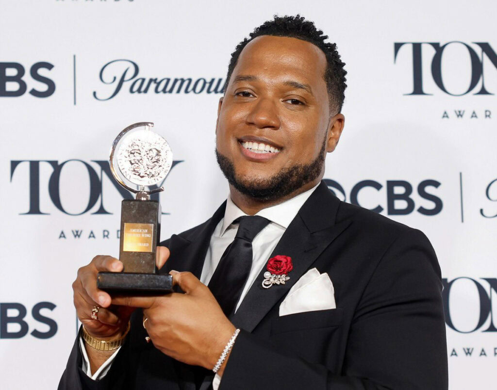 Jacobs-Jenkins in a black suit and tie, smiling while holding a Tony Award against a backdrop with logos. His expression conveys joy and pride.