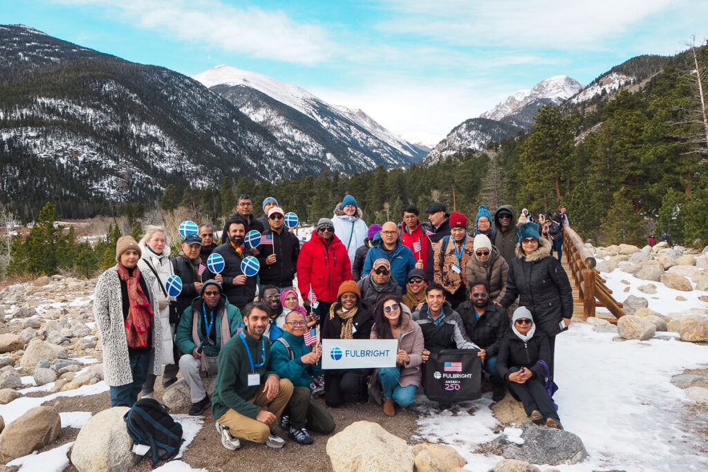 A group of people poses outdoors in a snowy mountain landscape in Rocky Mountain National Park. They hold Fulbright signs, expressing unity and happiness. Majestic peaks and pine trees provide a scenic backdrop.