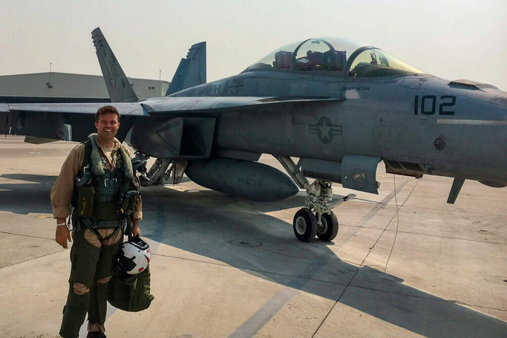 Jeff McLean standing in front of a fighter jet on a concrete airfield, wearing a pilot suit and carrying a helmet and gear