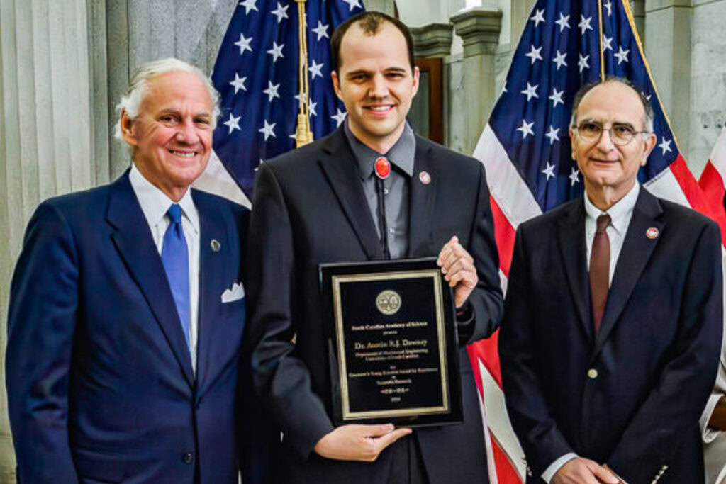 Austin Downey holding an award plaque next to the Governor of South Carolina and another man, all three wearing dark suits and ties, standing in front of two American flags