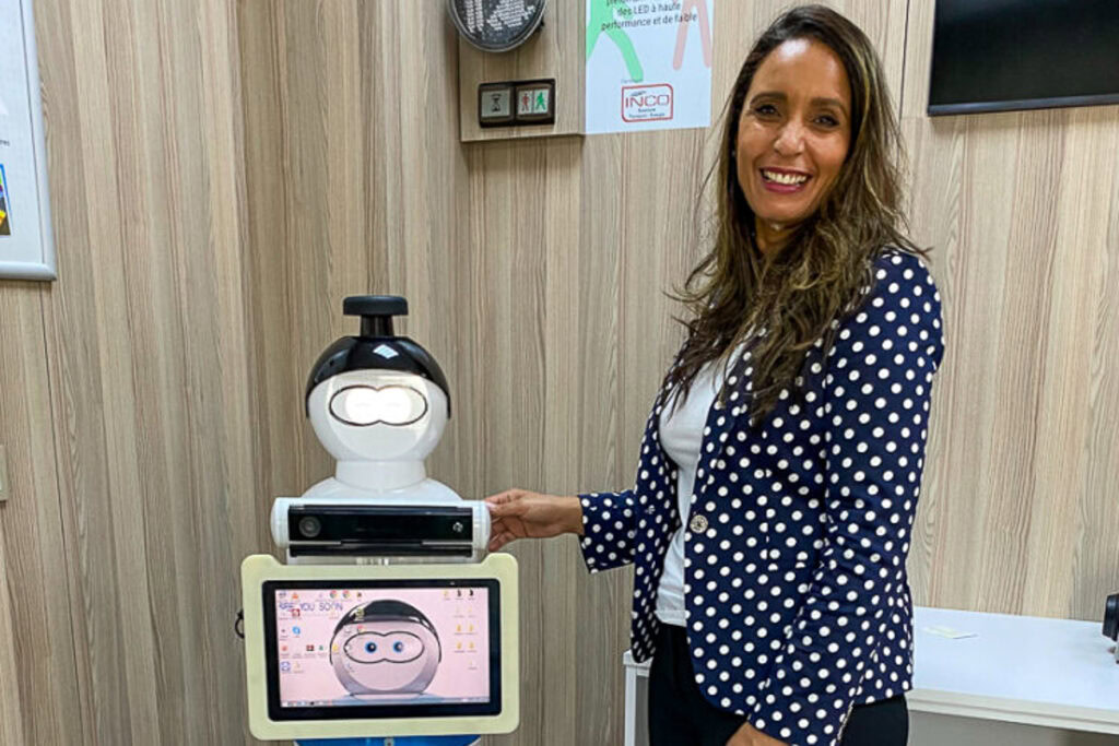 Rachida Parks wearing a navy blue polka dot blazer stands next to a small robot with a digital face in a medical office setting