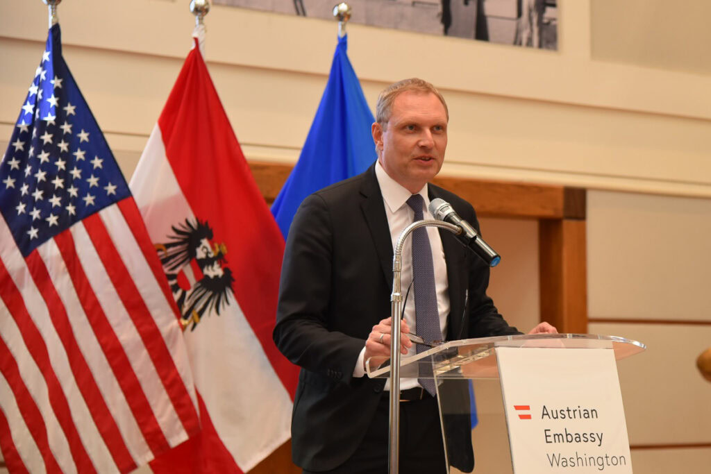 Man in dark suit and tie standing at podium speaking in front of American and Austrian flags