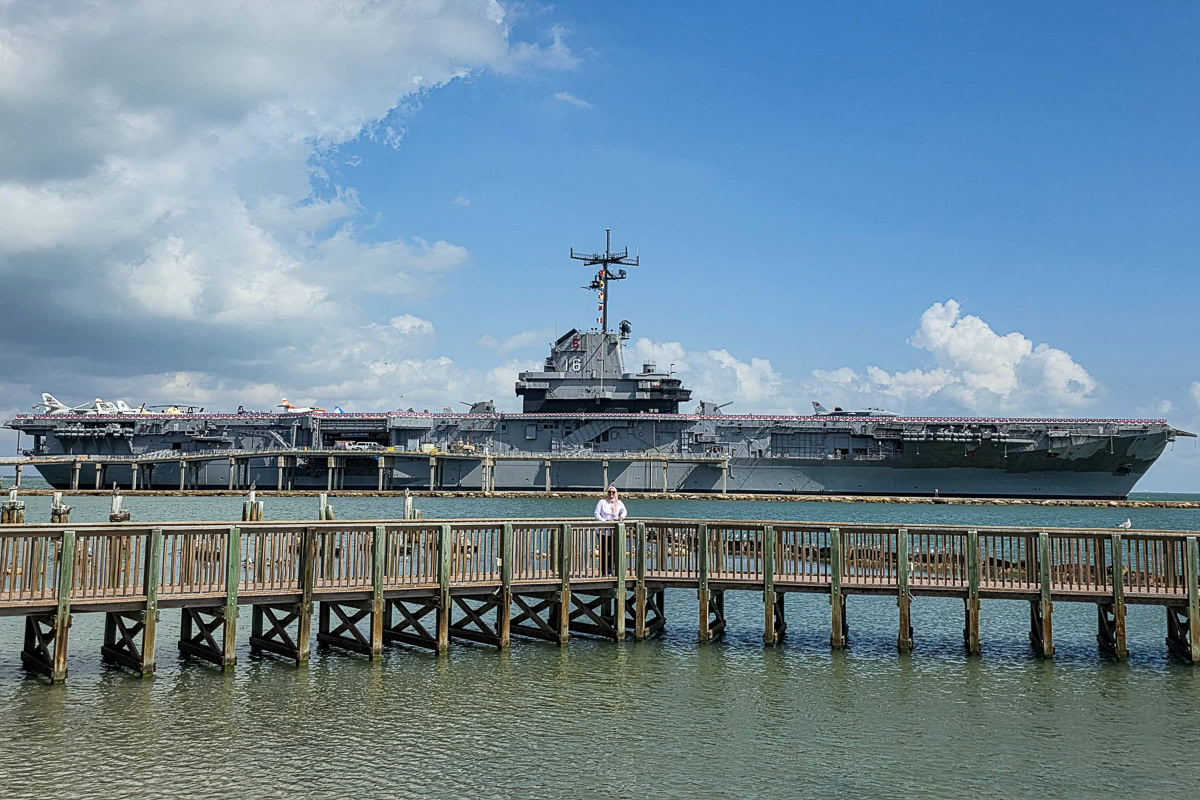 A large aircraft carrier sits in the water under a partly cloudy sky. In the foreground, a wooden pier extends out, with a person standing on it.