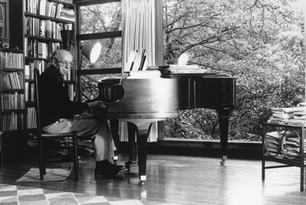 Aaron Copland seated at a grand piano in a room in his home with bookshelves and large windows showing trees outside
