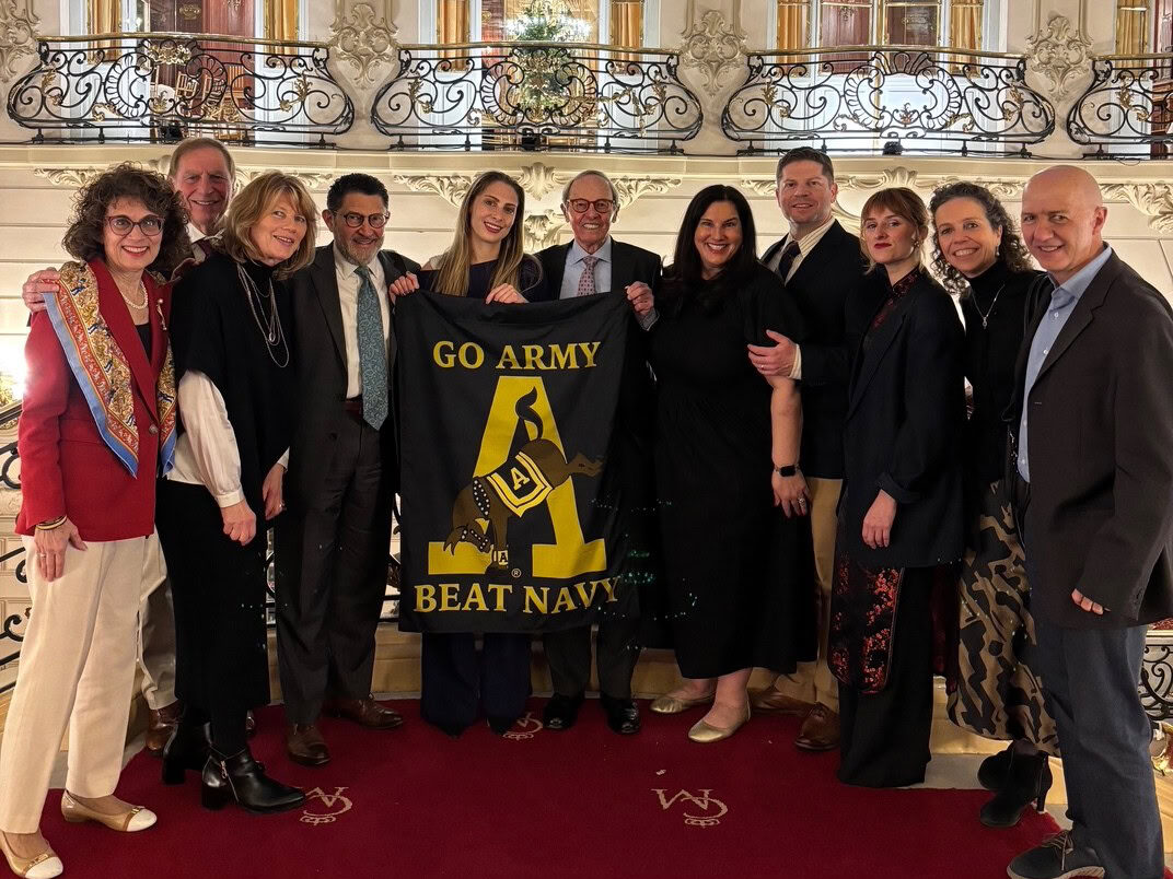 Boroff and group of people smile indoors, holding a "Go Army Beat Navy" banner. The ornate setting features elegant railings, exuding a celebratory tone.