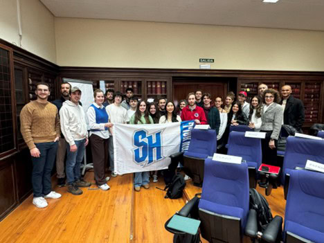 A group of American students in a Spanish university classroom holding a white flag with blue "SH" letters for Seton Hall, surrounded by blue chairs and dark wood bookshelves.