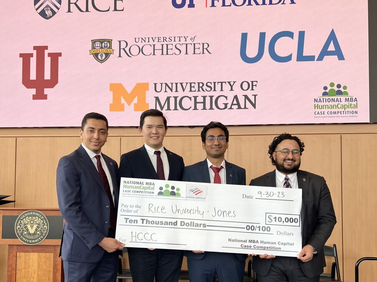 Four men in suits holding a large mockup of a check on a stage with university logos in the background. Text on the check says National MBA Human Capital Case Competition, Date: 9-30-23, 
Pay to the Order of: Rice University - Jones, Ten Thousand Dollars
$10,000