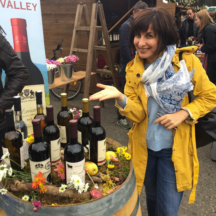 A market scene with a woman in a yellow coat and blue scarf smiling beside a display of wine bottles on a barrel and a wine marketing poster behind it