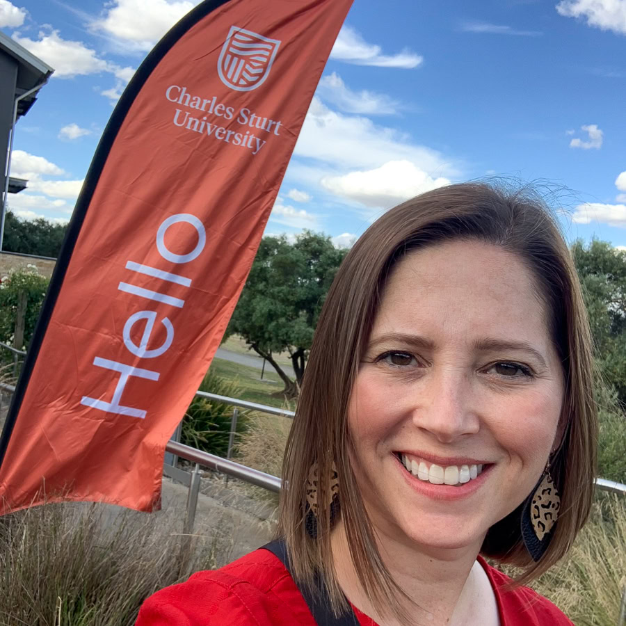 A smiling woman standing outdoors in front of a field in Australia, with a red banner featuring a university logo with the name Charles Sturt University and the word Hello.