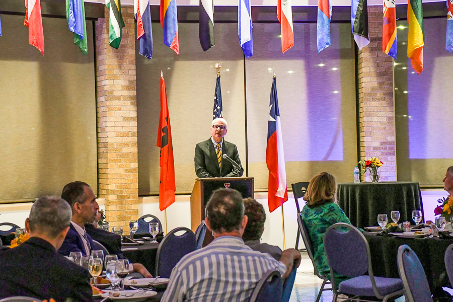 Texas Tech Provost Dr. Hendrick wearing a suit and tie standing at a podium with flags of the U.S. and Texas behind him and international flags hanging from ceiling, speaking to seated guests at round tables