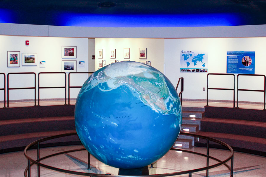 An indoor exhibit with a a large blue globe surrounded by a circle of metal railings in the center of a room with walls featuring a display of framed photos of international scenes alongside some informational posters with a Fulbright logo