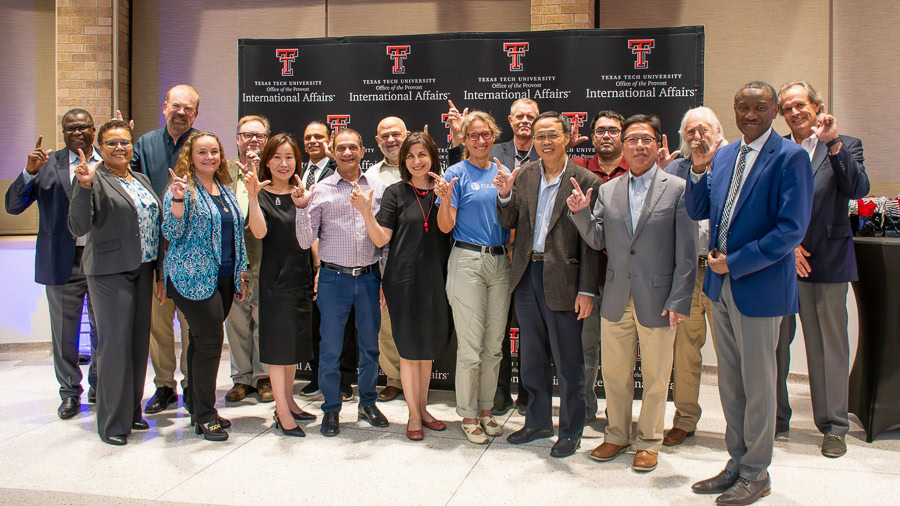 A group of around twenty people standing together in a posed arrangement. They are indoors, in front of a backdrop that features the Texas Tech University logo and the phrases Office of International Affairs.