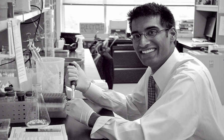 Reddi seated in a lab wearing a shirt and tie, holding a pipette, with lab equipment in front of him and a desk and office equipment in the background.