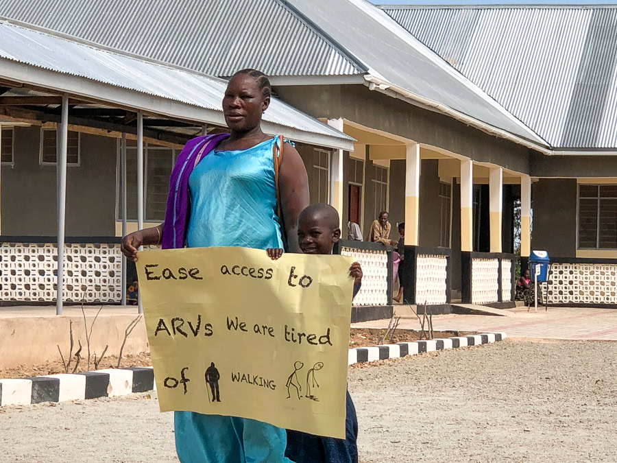 A woman and child hold a yellow sign in front of a building with metal roofing, columns, and a patterned railing. The sign says, "Ease access to ARVs We are tired of WALKING"