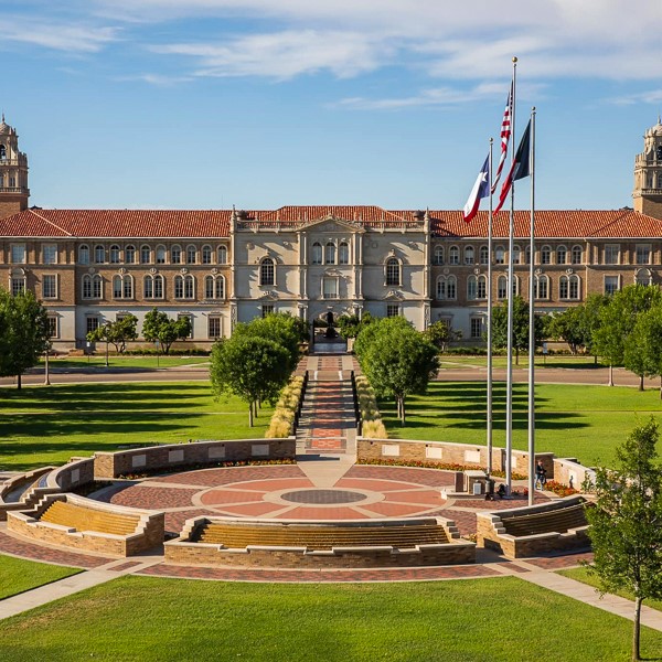 A grand campus building with a red-tiled roof stands behind a circular plaza with flagpoles, surrounded by green lawns and trees under a blue sky.