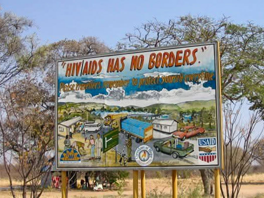 Billboard in a rural area in front of trees, with text "HIV/AIDS Has No Borders" depicting a travel scene. Text says, "HIV/AIDS HAS NO BORDERS. Please travellers remember to protect yourself every time. USAID."