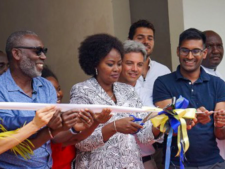 A group of people at a ribbon-cutting ceremony; a woman in the center cuts a white ribbon with blue and yellow bows while Reddi stands next to her and holds the ribbon.