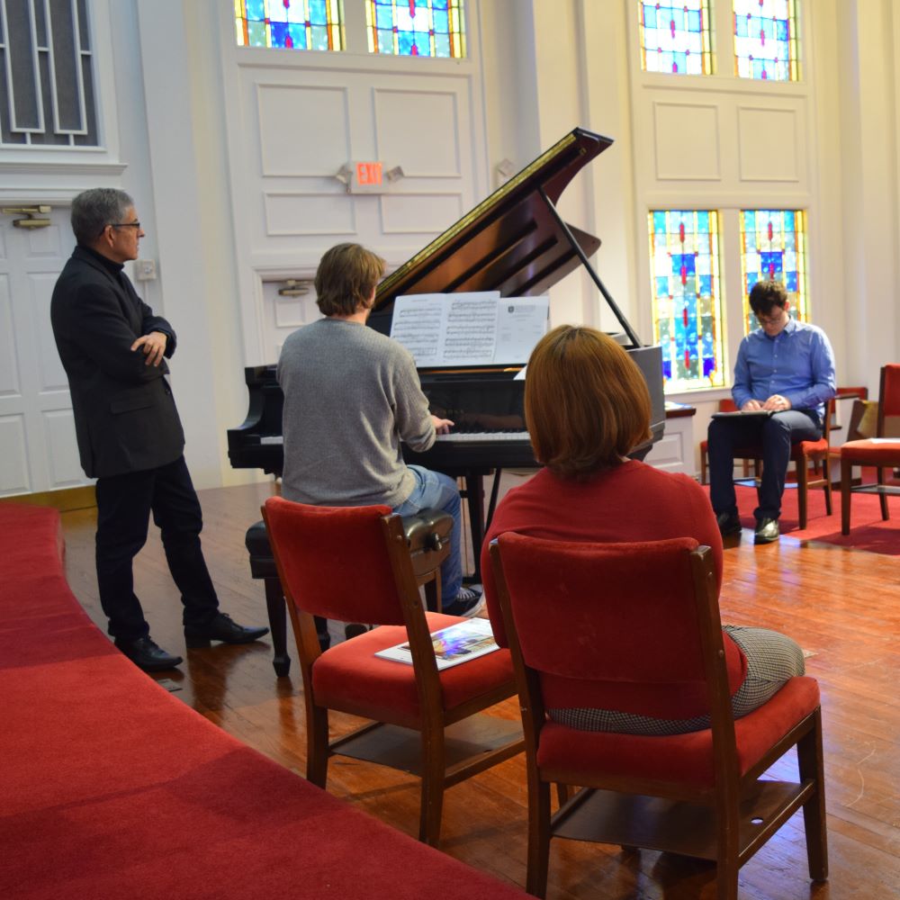 A pianist plays a grand piano in a well-lit room with stained glass windows. Three people watch intently, creating a focused and reflective atmosphere.