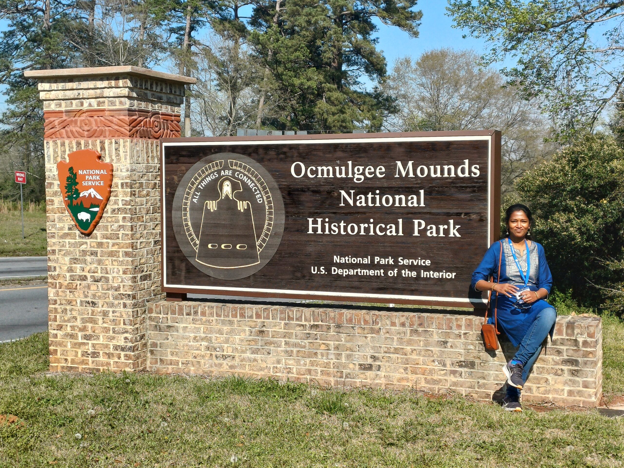 Woman in traditional Indian clothing sitting on brick wall in front of sign saying Ocmulgee Mounds National Historic Park