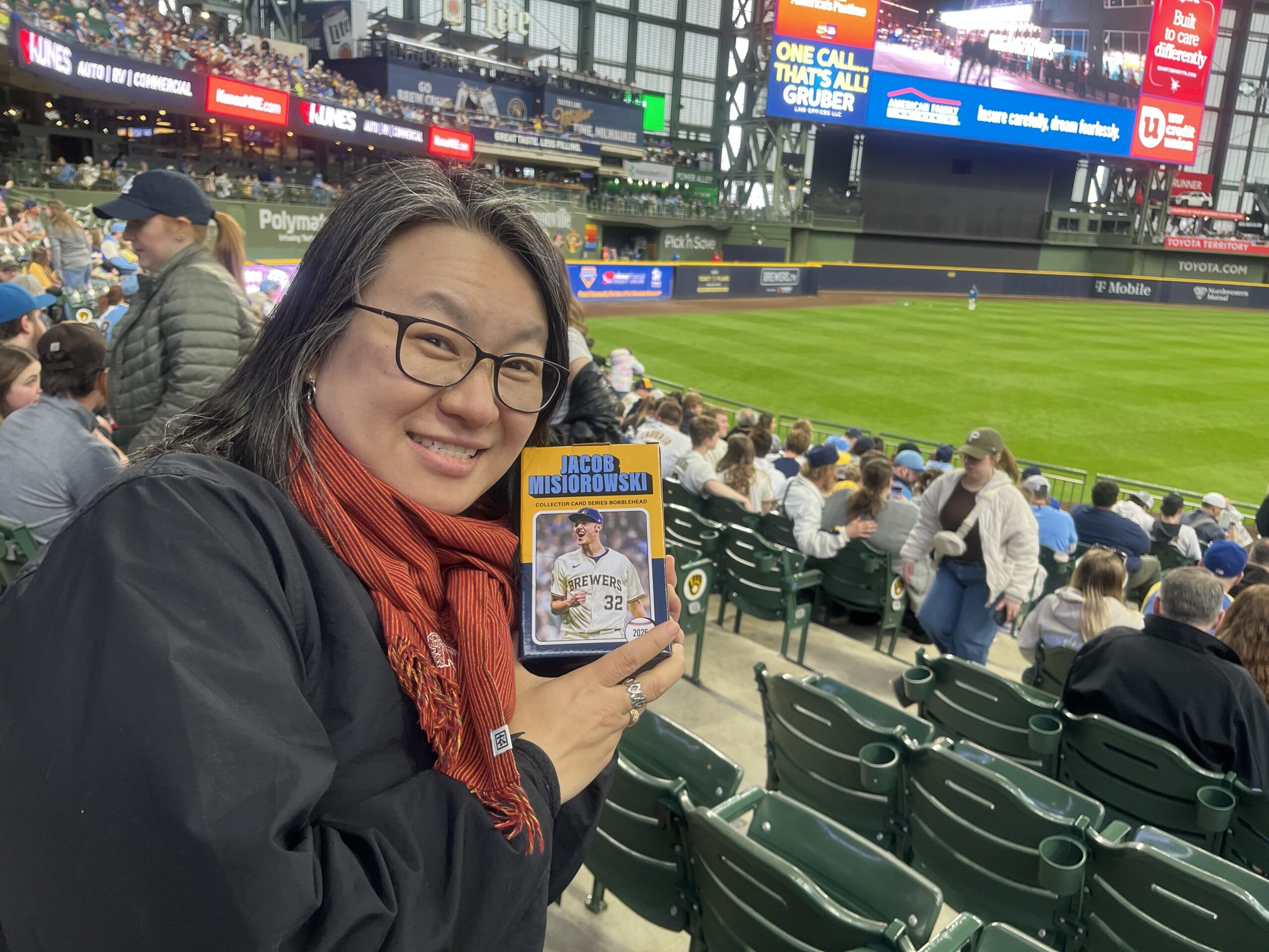 A woman in a black jacket and red scarf sitting in a green stadium seat at a baseball stadium, holding a baseball card with a player's image on it.