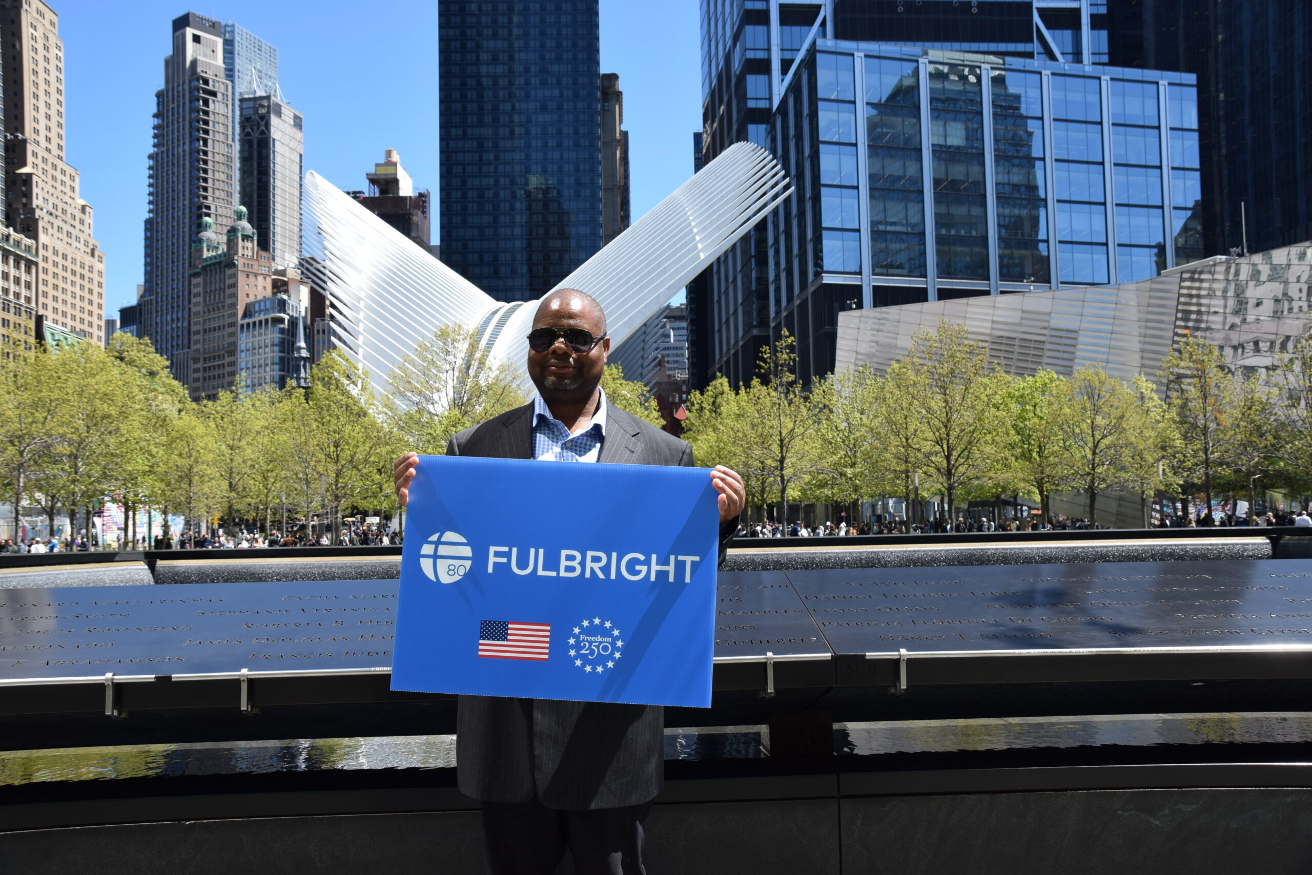 Man in suit standing in front of 9/11 Memorial Pool holding blue sign that says Fulbright with American Flag and Freedom 250 logo, with white wings of the Oculus building and a historic church steeple behind him