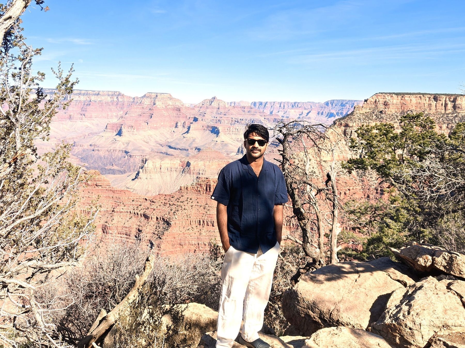 Man standing at the rim of the Grand Canyon, wearing a navy blue short sleeved shirt and white slacks, with some evergreen trees and leafless tree trunks near him and the red rocks of the canyon in the distance behind him.