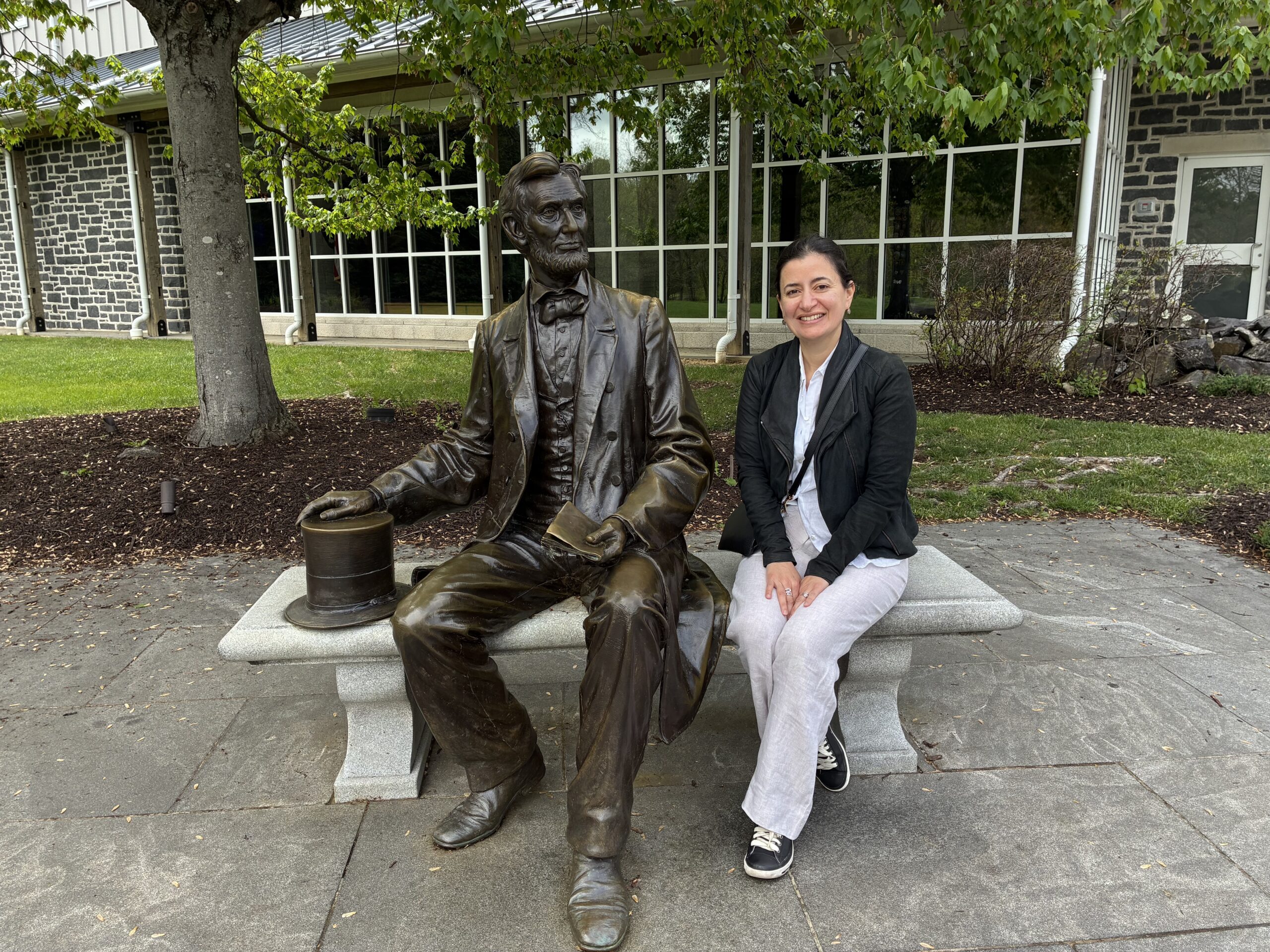 A woman sitting on a stone bench next to a bronze sculpture of Abraham Lincoln, posed sitting on the bench and holding his tophat.