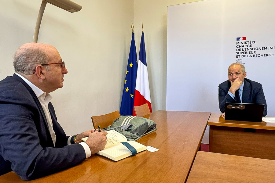 Two men sit at separate tables in a formal meeting room, one writing and the other pensive, with EU and French flags in the background.