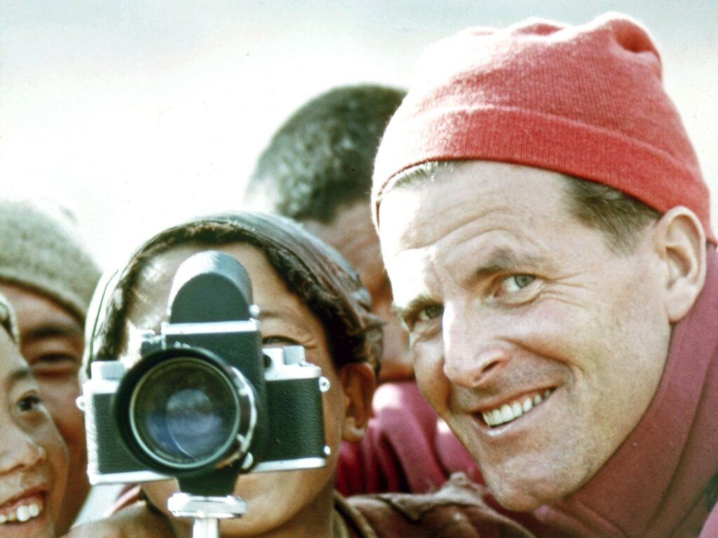 Man in red knit hat holding camera up for a young woman to look through with a group of people gathered around.