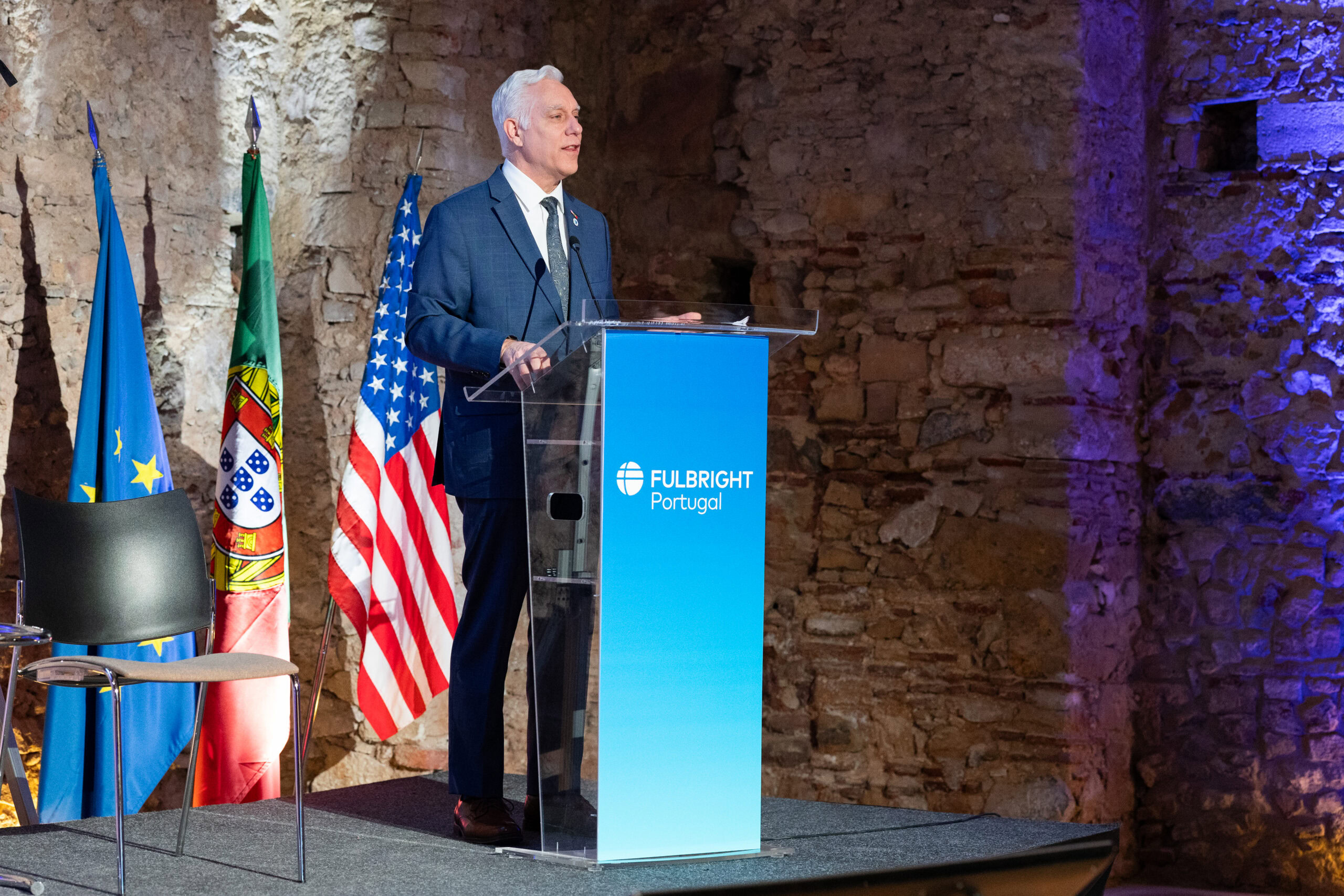 Man in suit standing at podium that says Fulbright Portugal, against the wall of a historic stone structure, with flags of Portugal and the United States behind him