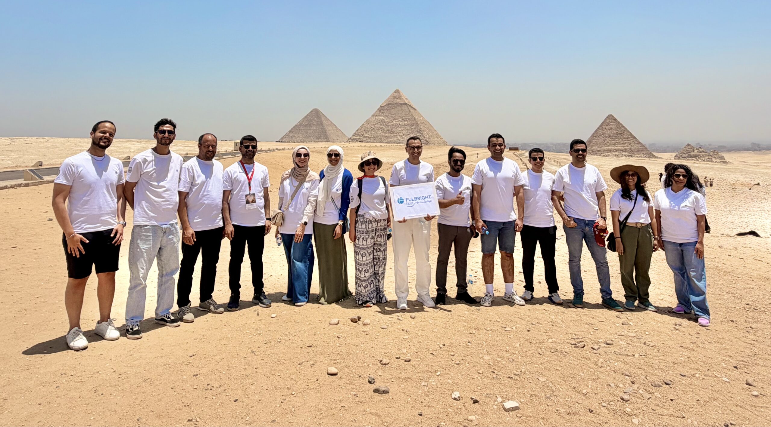 A group of 14 people, all wearing white shirts, standing in a row with Egyptian pyramids in the bakground. The man in the center of the row is holding a sign that says Fulbright.