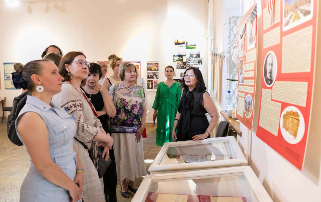 Group of about 10 people gathered around a museum exhibit case and looking up at a display poster on the wall with images of historic photos and documents on a red background.