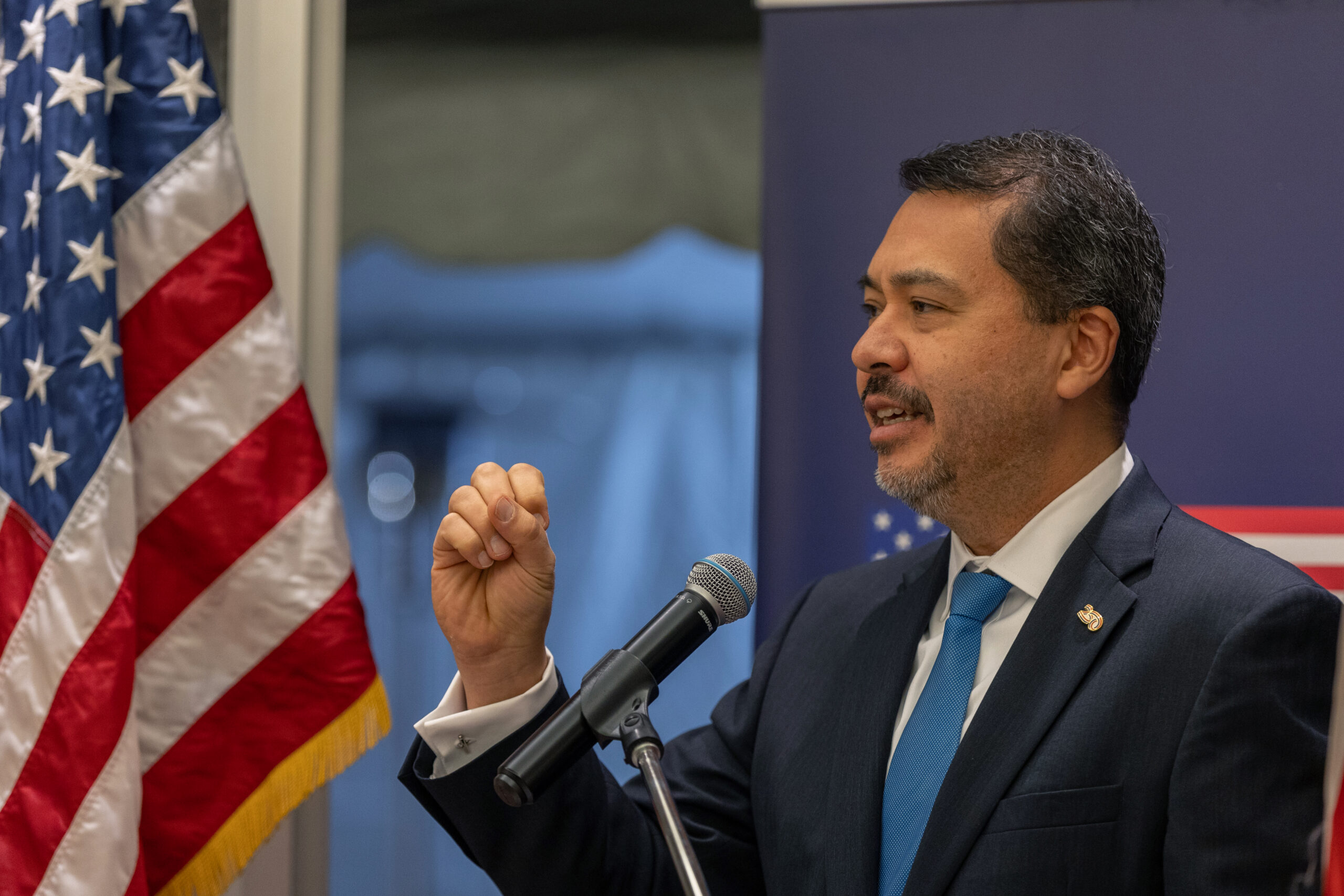 Man wearing a dark suit and blue tie and a lapel pin standing at microphone next to American flag,