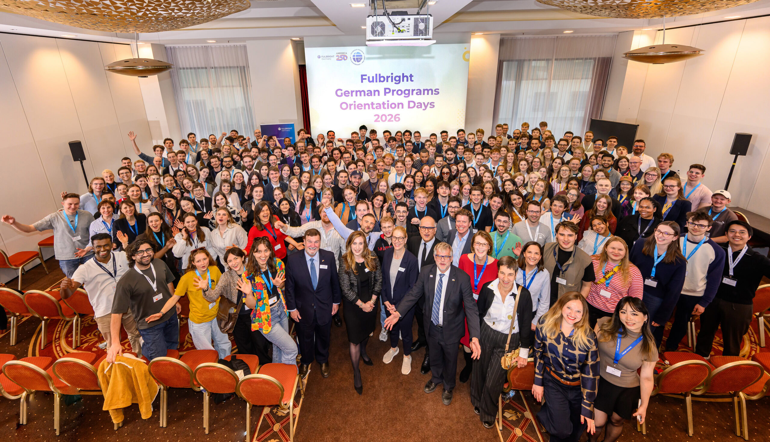 Group of 250 people wearing business attire standing in front of theater style chairs in a large meeting room with a screen behind them that says German Program Orientation Days 2026.