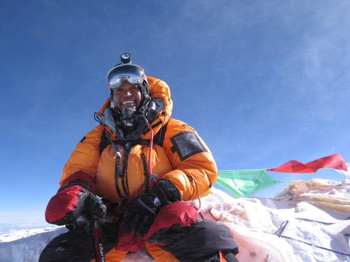 Woman at the peak of Mt. Everest wearing orange parka and mountaineering gear, with prayer flags in background