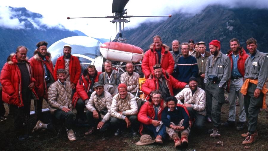 A group of men  wearing parkas and mountaineering gear sitting and standing in front of a helicopter with a mountain peak and clouds behind them