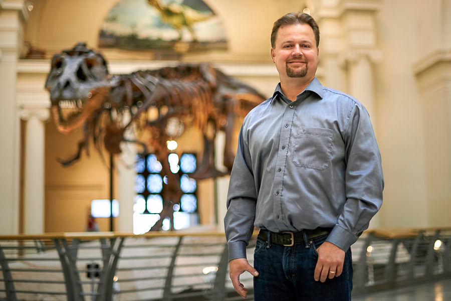 A man in a blue button down shirt stands in front of a T. rex skeleton displayed in the atrium of the Field Museum of Natural History 