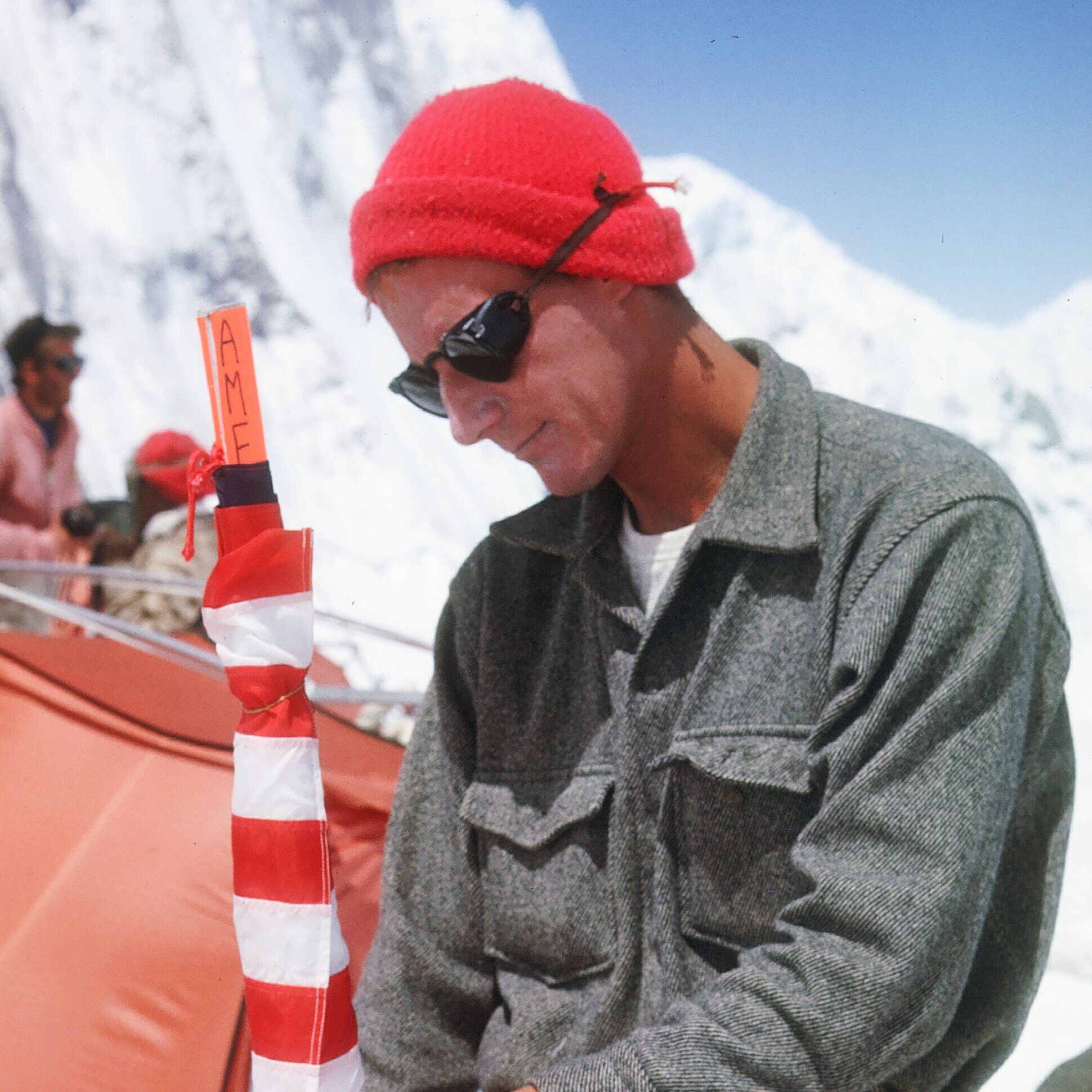 Climber standing in front of orange tent and mountain peak, packing rolled up American flag in preparation for his ascent to the summit of Mt. Everest
